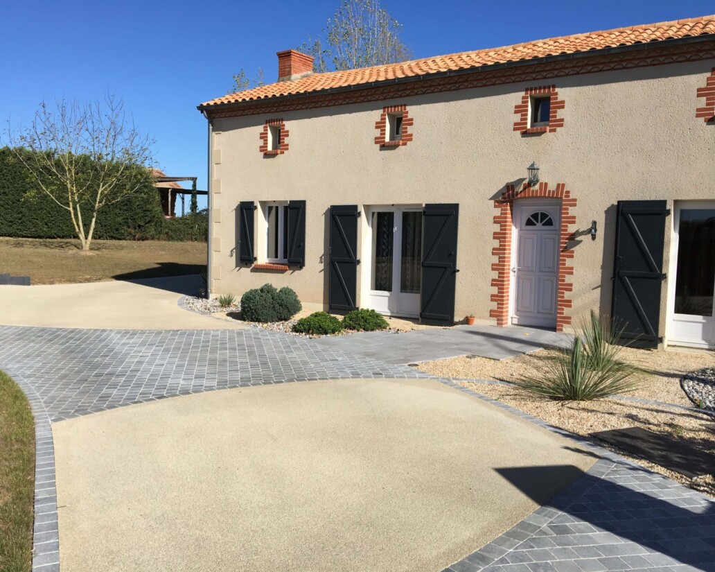 all&eacute;e d'entr&eacute;e en b&eacute;ton drainant beige & pav&eacute;s gr&egrave;s gris. La maison est en enduit beige, contours des ouvertures en briques rouges